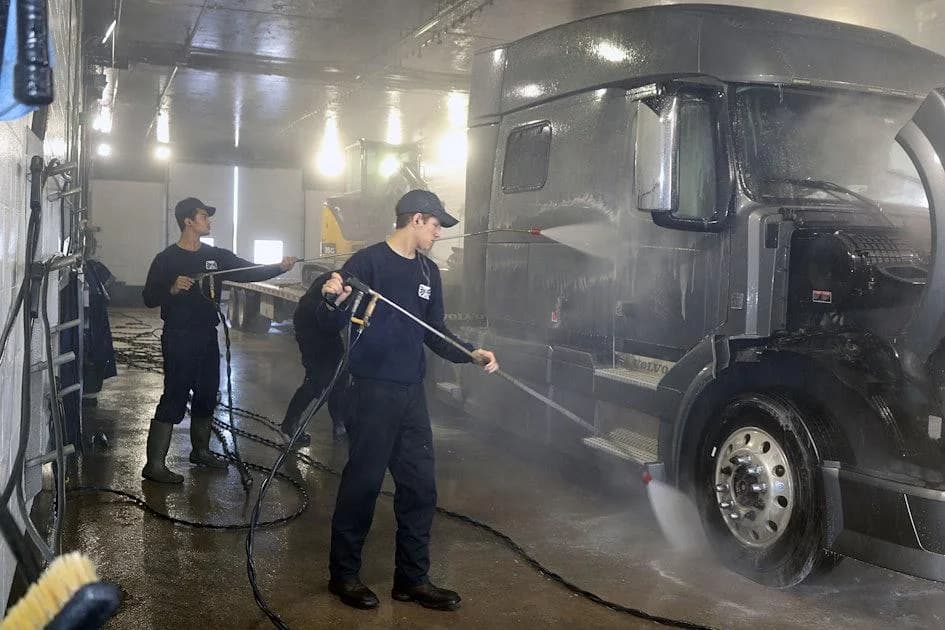 Technicians pressure-cleaning a dark semi-truck from roofline to wheels.